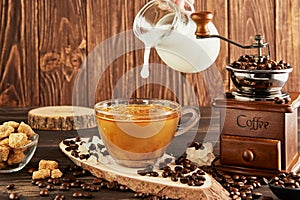 Pouring coffee from glass jug milk into glass cup with coffee, vintage coffee grinder and lump sugar on wooden