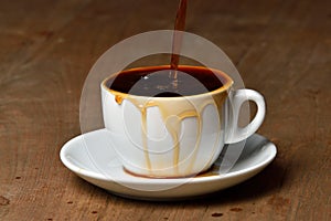 Pouring coffee into overflowed white porcelain mug with saucer. Cup with overflowing coffee. Full cup of coffee on wooden table