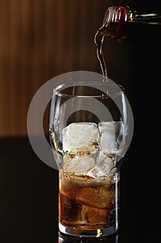 Pouring cola from bottle into glass with ice cubes on table