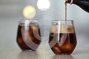 Pouring cola from bottle into glass with ice cubes on table against blurred background.