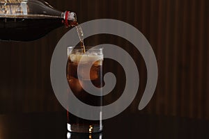 Pouring cola from bottle into glass with ice cubes on table against blurred background