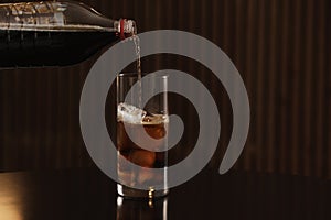 Pouring cola from bottle into glass with ice cubes on table against blurred background.