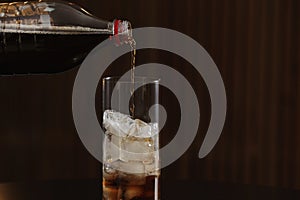 Pouring cola from bottle into glass with ice cubes on table against blurred background