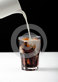 Pouring fresh milk into glass of black coffee with ice cubes on white board and black backdrop