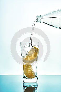 Pouring fresh water in glass with lemon from plastic bottle on blue background. Fresh drinking water glass with bubbles blue backg