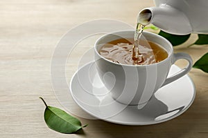 Pouring green tea into white cup with saucer and leaves on wooden table, closeup. Space for text