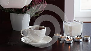 Pouring hot black tea in white ceramic cup with lighted scented candles around and flowers slow living concept real time