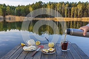 Pouring hot tea into a glass mug from a thermos in the morning next to the lake and forest. Breakfast on a wooden table