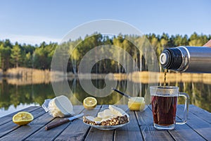 Pouring hot tea into a glass mug from a thermos in the morning next to the lake and forest. Breakfast on a wooden table