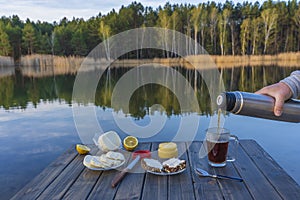 Pouring hot tea into a glass mug from a thermos in the morning next to the lake and forest. Breakfast on a wooden table