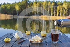 Pouring hot tea into a glass mug from a thermos in the morning next to the lake and forest. Breakfast on a wooden table