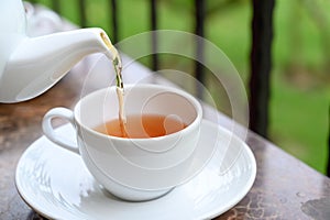 Pouring hot tea from white ceramic teapot into porcelain cup. Outdoor breakfast on balcony cafe