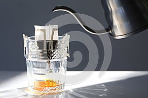 Pouring hot water into glass with drip coffee bag from kettle on light grey table, closeup