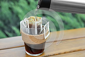 Pouring hot water into glass with drip coffee bag from thermos on wooden table, closeup