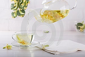 Pouring linden tea from teapot into transparent cup. Glass cup and tea pot with herbal lime tree tea on white background.