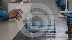 Pouring liquid chemical from a plastic wash bottle dispenser to test tubes on a rack on a black granite slab in