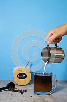 Pouring milk into black coffee in a glass, stevia in the background