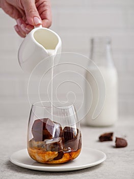 Pouring milk into a glass of espresso coffee with frozen coffee cubes on bright background