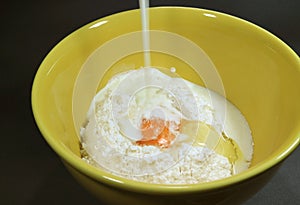 Pouring milk into yellow ceramic mixing bowl with dry flour and egg for making pancakes