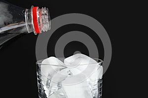 Pouring refreshing cola from bottle into glass with ice cubes on black background