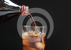 Pouring refreshing cola from bottle into glass with ice cubes on black background