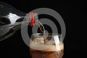 Pouring refreshing cola from bottle into glass with ice cubes on black background