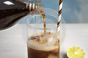 Pouring refreshing soda drink into glass with ice cubes and straw on table