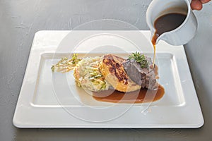 Pouring sauce on a dish from roast venison, flat bread dumpling and round shaped cabbage vegetable on a white plate, preparation