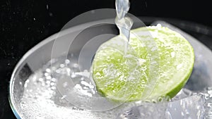 Pouring soda into a cocktail glass with lime and ice in slow motion on dark background, Refreshing cold cocktail