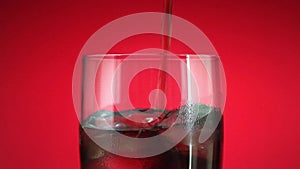 Pouring soft drink in glass with ice splash on red background. Cola or refreshment.