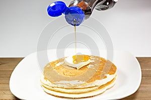 Pouring syrup from a blue tip bottle onto the square of butter on a stack of golden pancakes waiting to be eaten