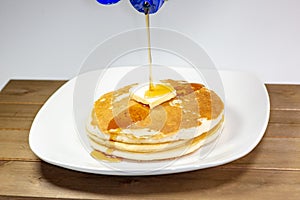 Pouring syrup from a blue tip bottle onto the square of butter on a stack of golden pancakes waiting to be eaten