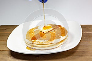 Pouring syrup from a blue tip bottle onto the square of butter on a stack of golden pancakes waiting to be eaten