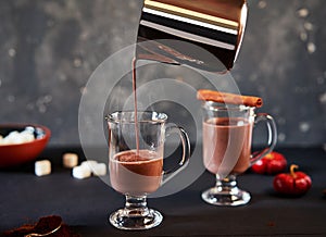 Pouring tasty hot chocolate cocoa drink into glass mug with ingredients on black table.