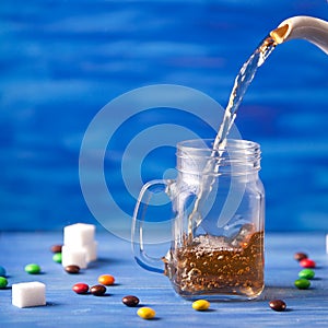 Pouring tea on blue background on blue wooden table with sugar and sweets. Drink flows from glass jar. Tea is pouring from a jug