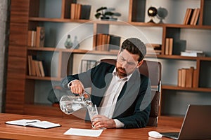 Pouring water, feeling tired. Businessman in formal clothes is working indoors