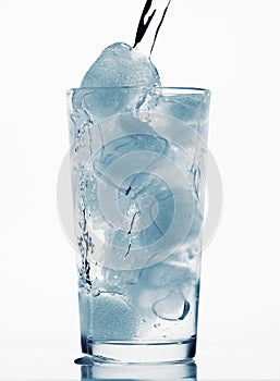 Pouring water in a glass full of ice cubes, white background, blue toned object