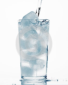 Pouring water in a glass full of ice cubes, white background, blue toned object