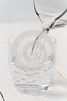 Pouring water from a glass jug into a glass. White wood background.