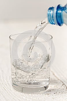 Pouring water from a plastic bottle into a glass. White wood background.