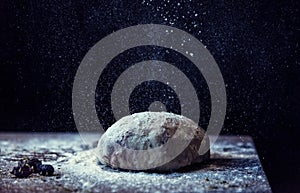 Pouring white wheat flour on a dough in a kitchen in front of black background, close up