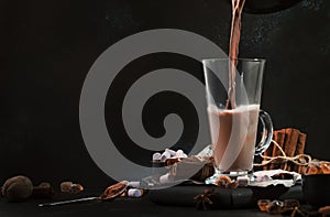 Pouring winter hot chocolate cocoa drink into tall glass with ispices on black table. Dark blue background. Copy space