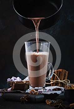 Pouring winter hot chocolate cocoa drink into tall glass with ispices on black table. Dark blue background. Copy space