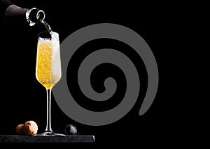 Pouring yellow champagne from bottle to glass with cork and wire cage on black marble board on black background. Space for text