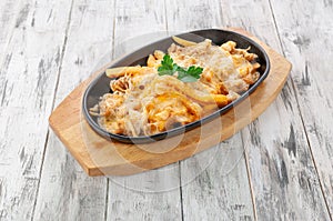 Poutine with chicken and french fries. Baked with cheese sauce. In a frying pan on a stand. Light wooden background. View from