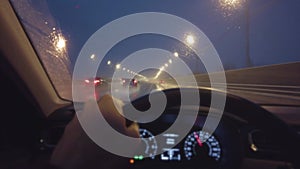 POV car driving shot in the rain at night. Driver's hand on a steering wheel