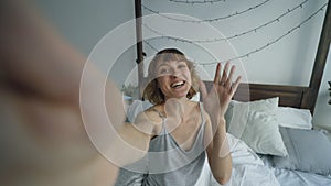 POV of cheerful woman having online video chat with friends using smartphone camera while sitting on bed at home