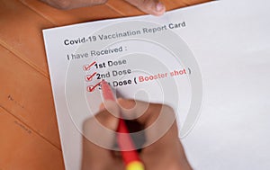 POV close up shot of hands checking Covid-19 vaccine report card and ticking 3rd or booster dose after vaccination