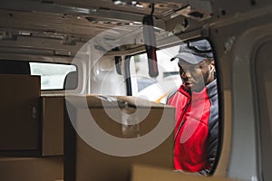 POV inside delivery van. Tired focused Black male courier in red jacket, black hat, and wireless headphones choosing the