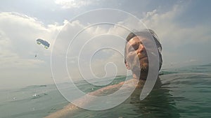 POV of man with beard swimming in sea water on sunny day Paragliding on background Summertime